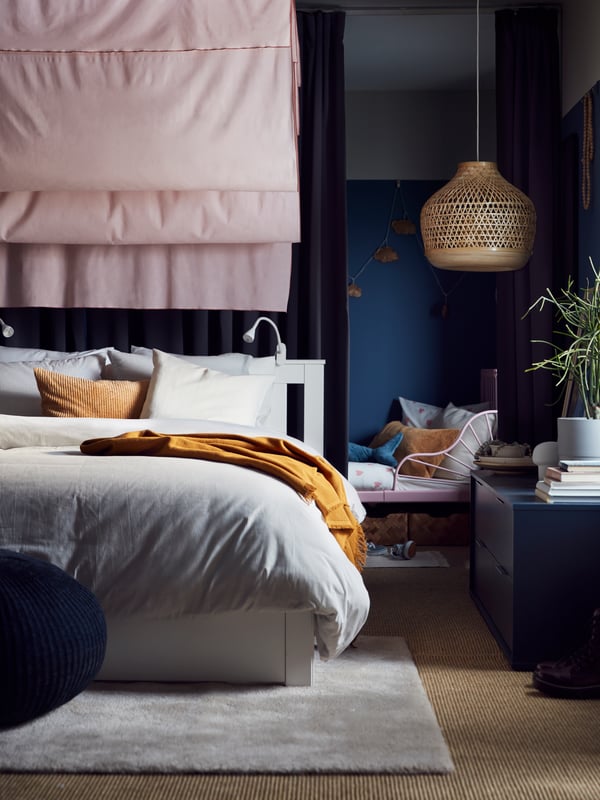 A white SONGESAND bed with gray striped bed linen, on an off-white rug and with pink curtains and a bamboo pendant above.