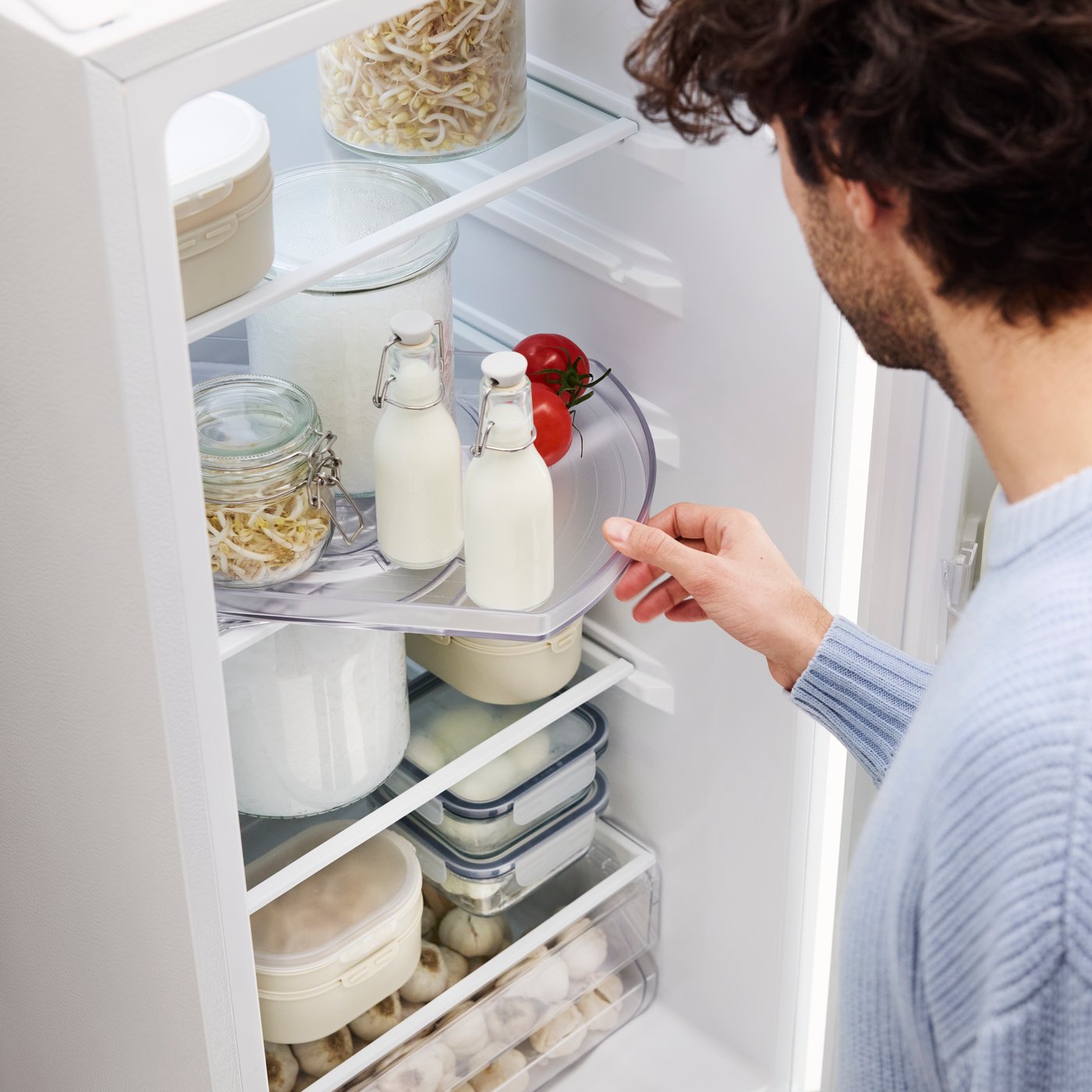 A person opens a fridge and pulls out a SNURRAD turntable, containing bottles of milk, leftovers in a jar and fresh tomatoes.