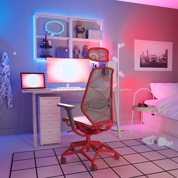 Un fauteuil gamer STYRSPEL gris et rouge devant des écrans allumés posés sur un bureau dans une pièce éclairée d’une douce lueur rouge.