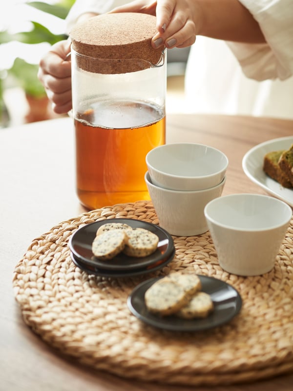 Un jarro IKEA 365+, tazas KRUSTAD de té y dos platos pequeños con galletas sobre un individual redondo SOARÉ, dispuesto sobre una mesa de madera. 