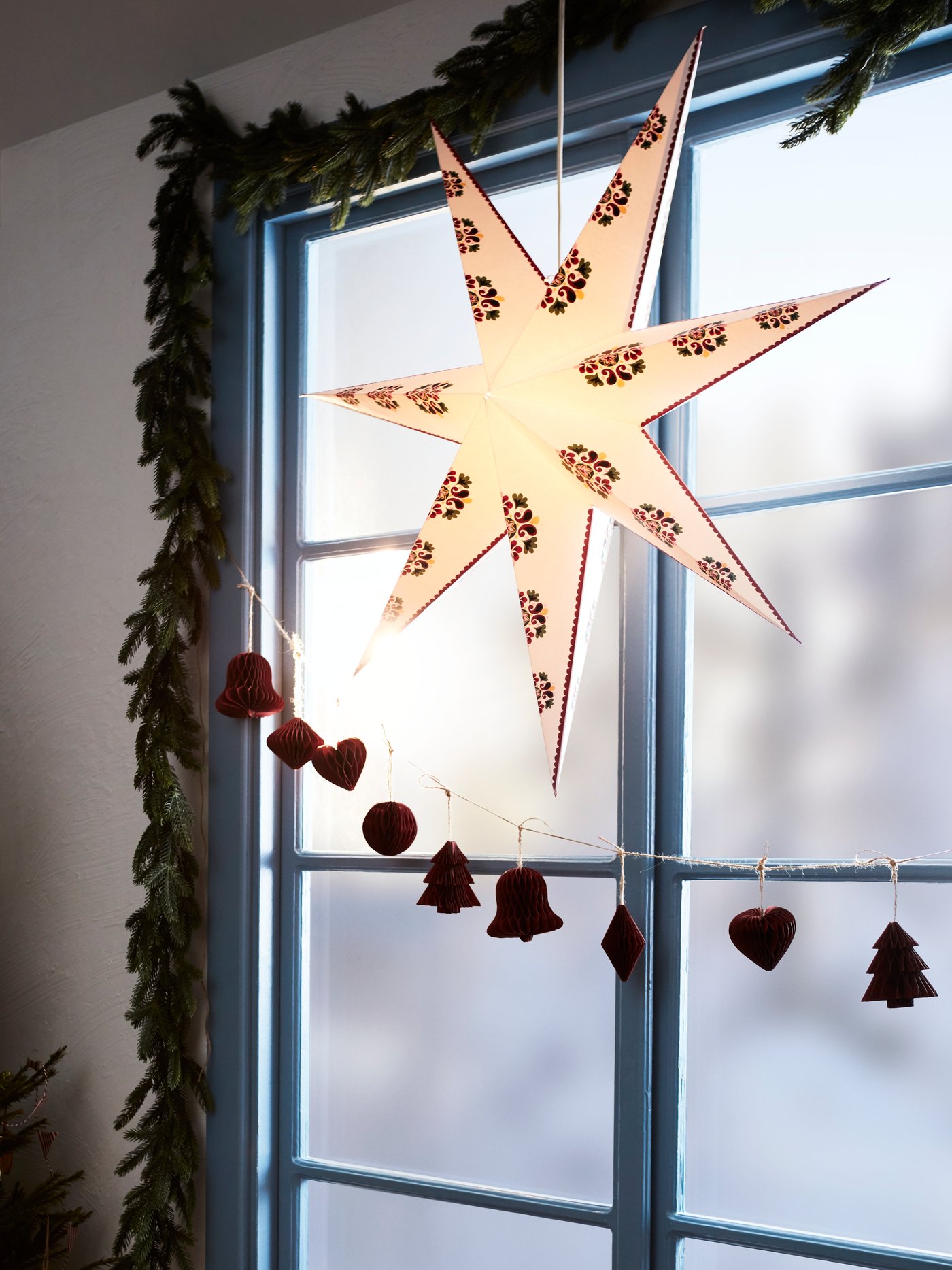 A STRÅLA star-shaped lampshade hangs in a window with VINTERFINT hanging decorations. A STRÅLA LED garland frames the window.