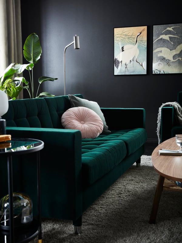 Canapé LANDSKRONA en velours vert avec coussin rose sur l’assise, et grande plante derrière lui, près d’un mur gris foncé orné d’images.