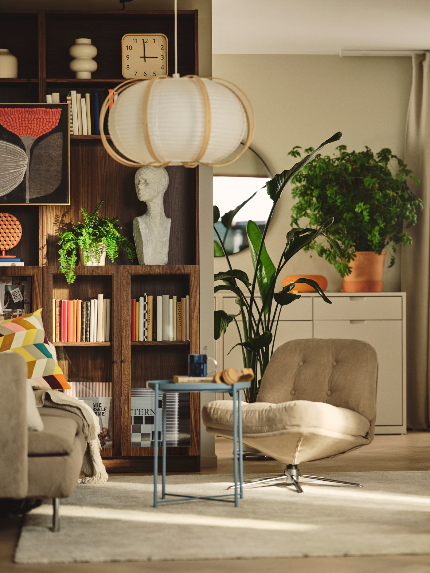 A living room features a brown BILLY bookcase with a walnut effect, filled with books beside plants and a soft chair.