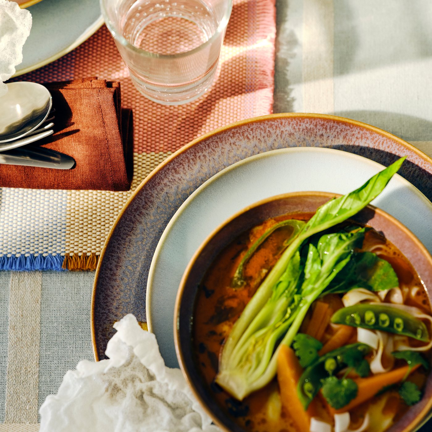 A table set with FÄRGKLAR bowls and plates filled with diverse food, a glass and tableware on a table mat.