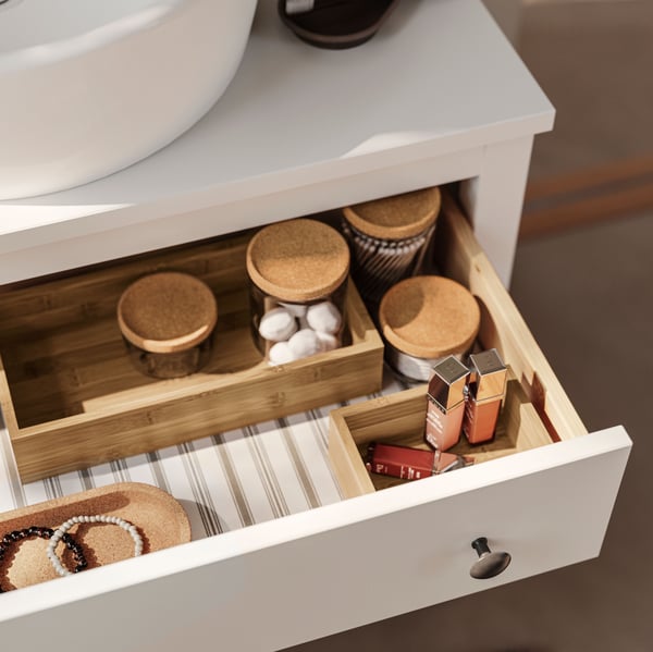 حامل حوض HEMNES أبيض به درج مفتوح داخل مرطبانات زجاجية بأغطية من الفلين وصواني TAVELÅN ومنتجات عناية.