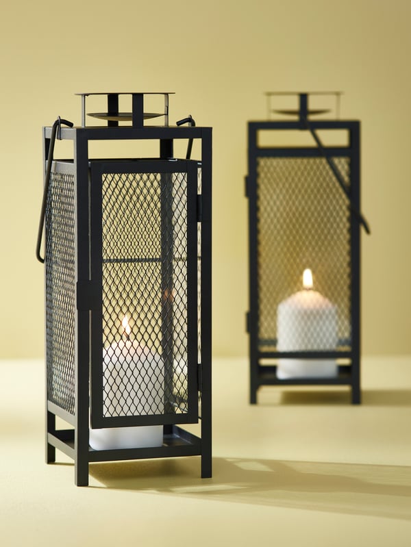 Two anthracite-coloured BERGGRAN lanterns, with lit candles inside, are sitting in a space with yellow walls and floors.