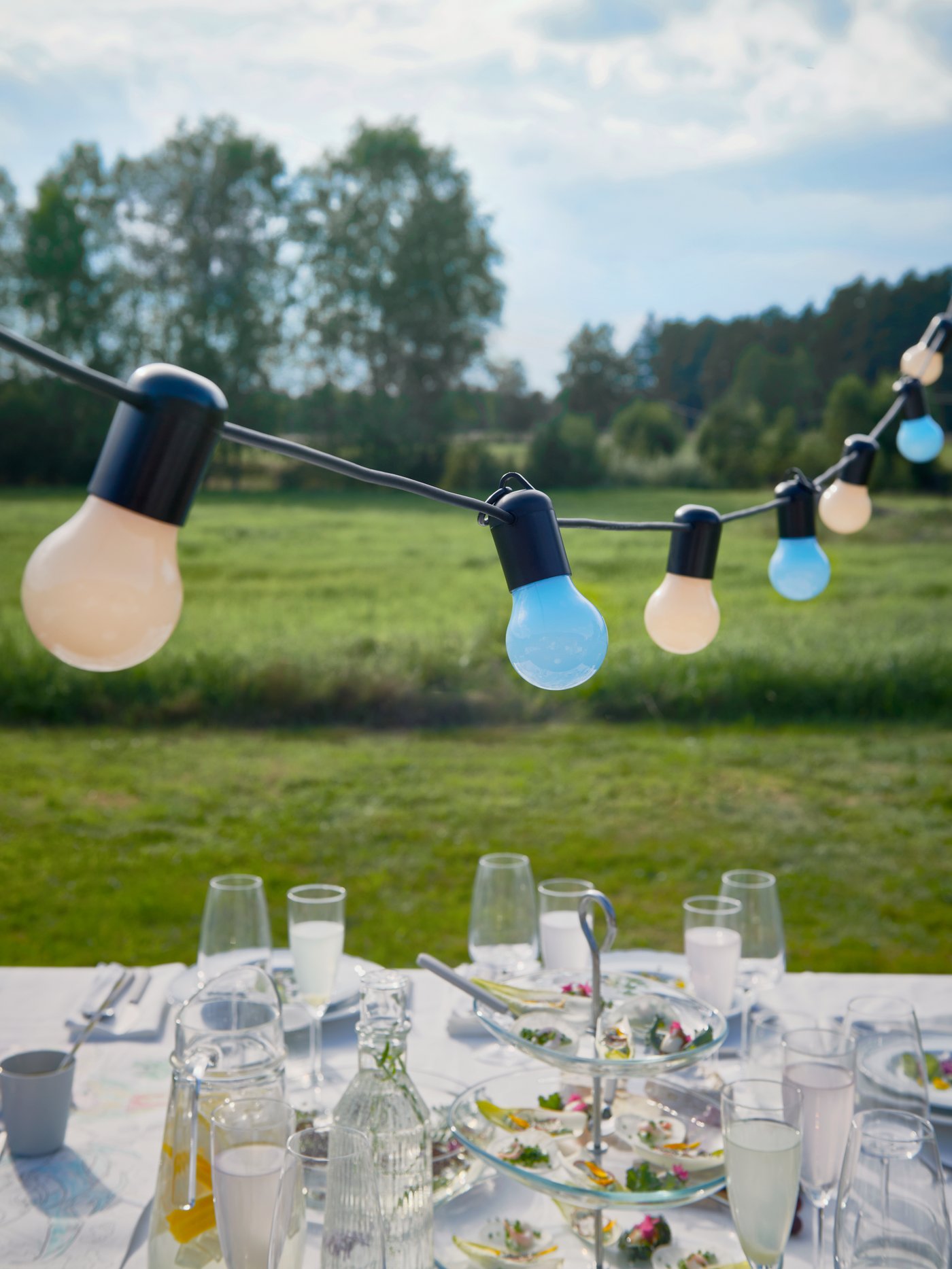 Eine bunte SOLVINDEN LED-Lichterkette hängt über einem Tisch, der für eine Hochzeitsfeier im Freien gedeckt ist.