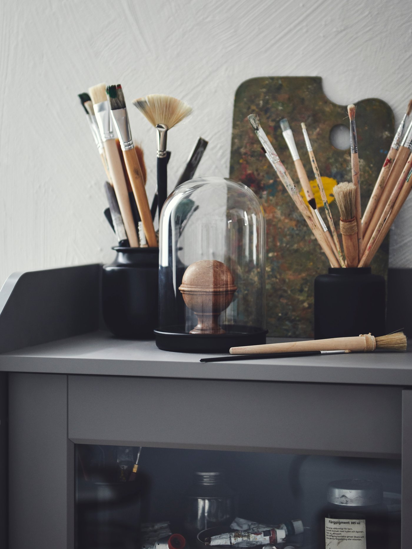 Sprawling bouquets of thin paintbrushes placed in a black KONSTFULL vase and a black jar atop a HAUGA cabinet.