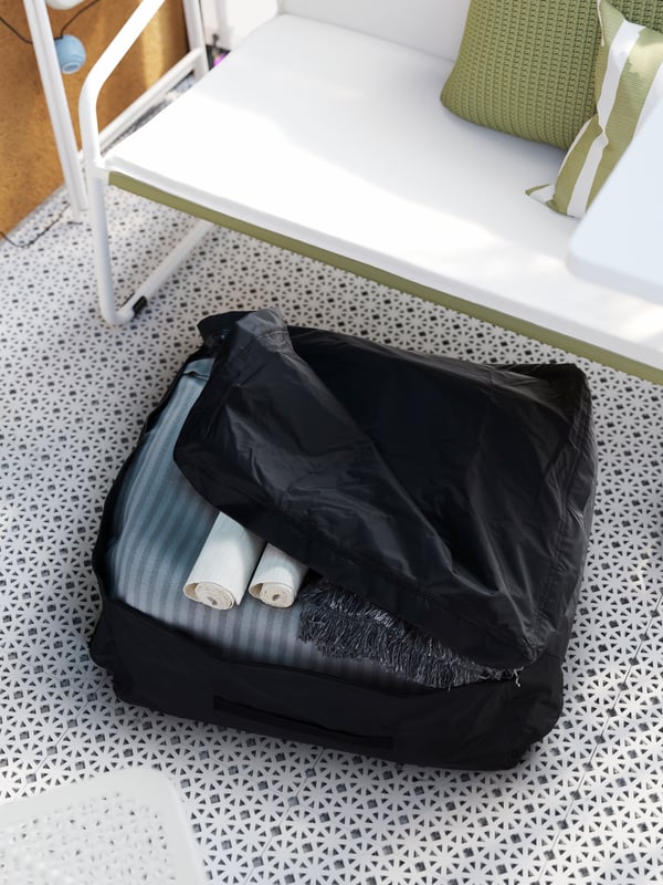 Una funda TOSTERÖ negra con mantas y textiles enrollados en su interior. Está posada sobre el suelo ALTAPPEN de una terraza y situada bajo un sillón INGMARSÖ blanco.