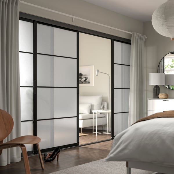 Eine SKYTTA Schiebetüre teilt ein Schlafzimmer vom Wohnzimmer. Beide Räume sind mit hellen Möbeln eingerichtet.