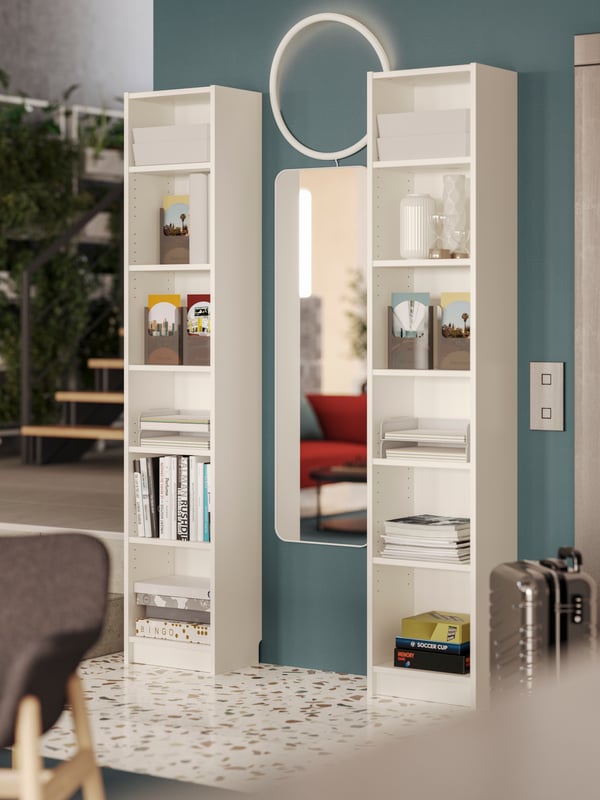 Two white BILLY bookcases filled with tourist brochures and magazines frame a mirror next to an elevator in a hotel lobby.
