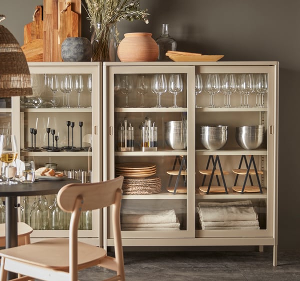 Beige IDÅSEN cabinet with sliding glass doors filled with serve ware in a dining room.