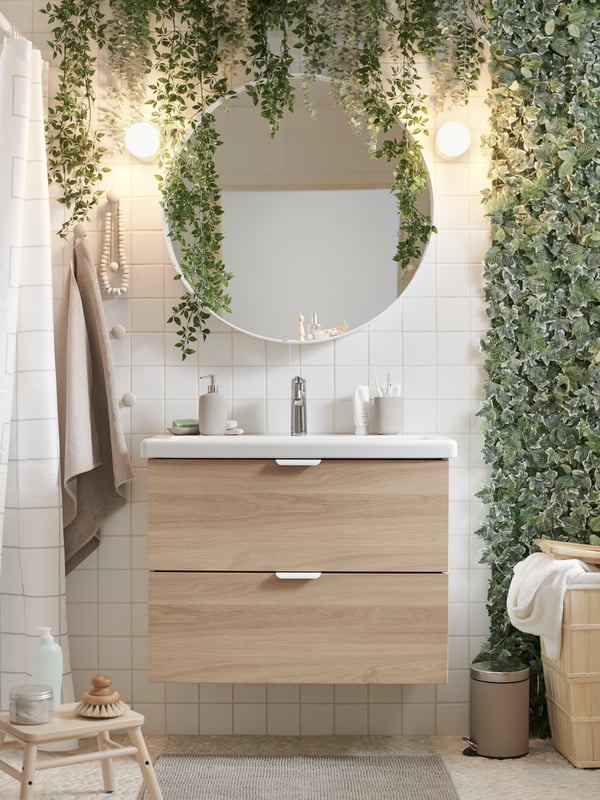 A bathroom decorated with FEJKA artificial plants, an ENHET/TVÄLLEN wash-stand, a VILTO birch step stool and a round mirror.