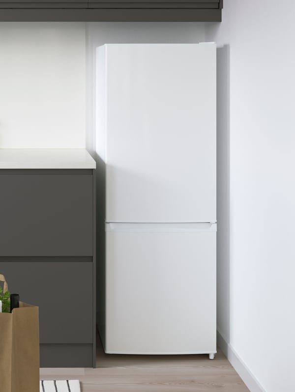 A freestanding LAGAN fridge/freezer combination in white in the corner of a kitchen with white walls and grey storage.