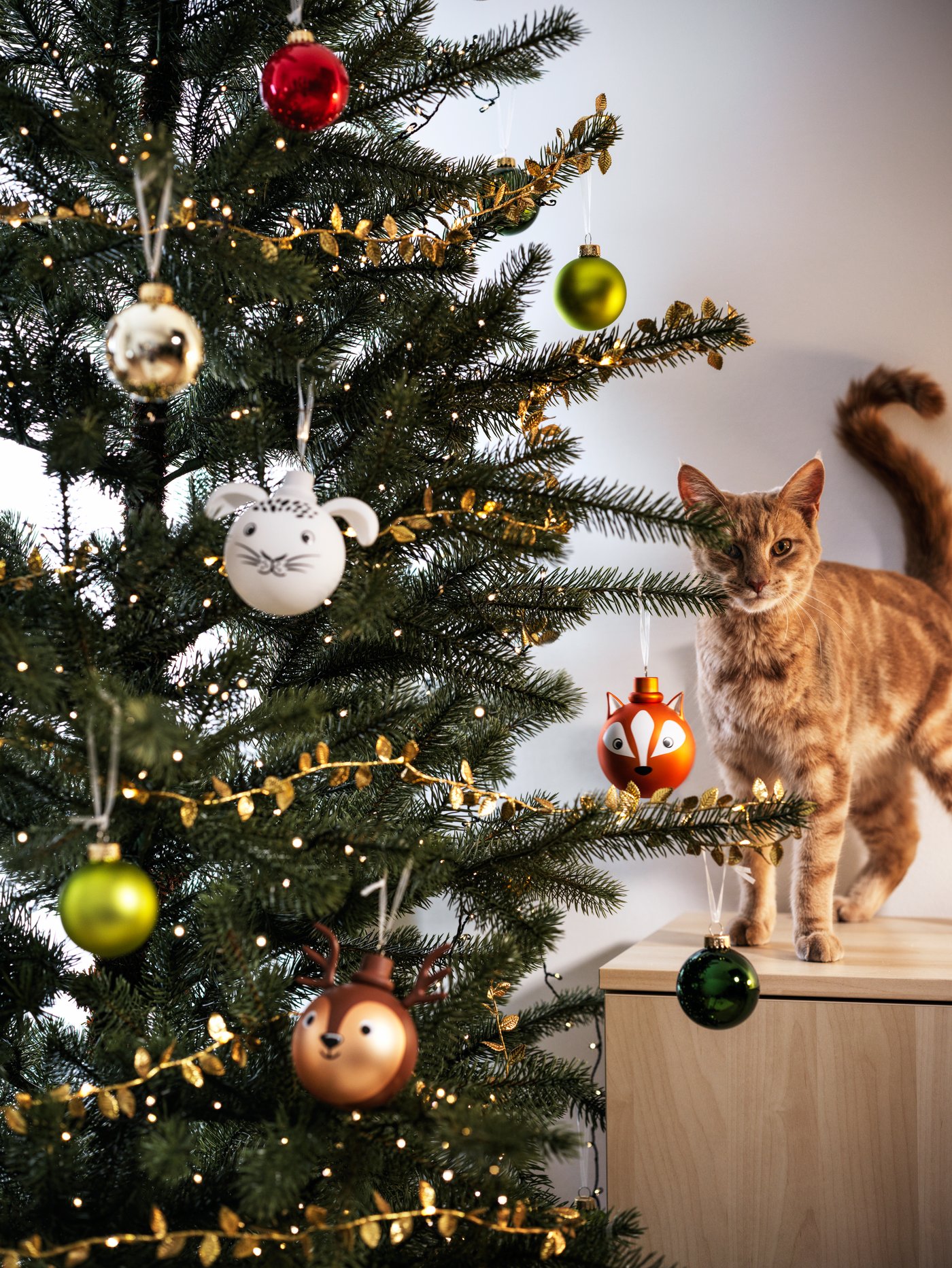 Un arból de navidad con decoraciones de animal al costado de un árbol. Al costado hay un gato sobre el mueble.