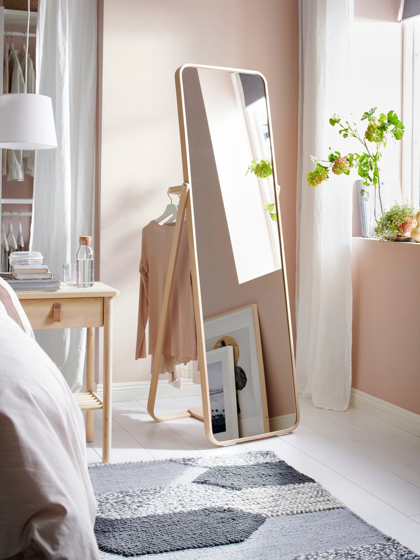 IKORNNES standing mirror featuring clothes hanging neatly at the back in the corner of a bedroom next to the windows. A BJORKSNAS bedside table stands next to the mirror before a grey rug with patterns. 