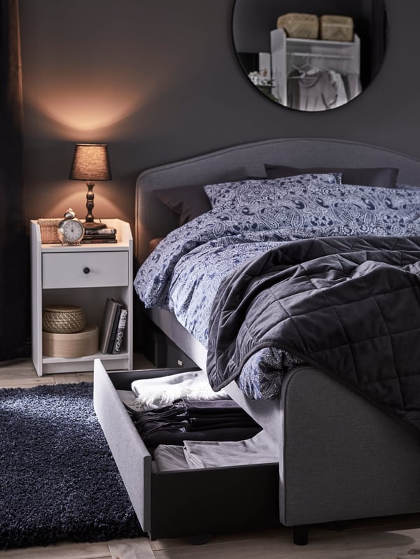 A grey HAUGA upholstered bed with the storage drawer open, and a white bedside table beside it holding a small table lamp.