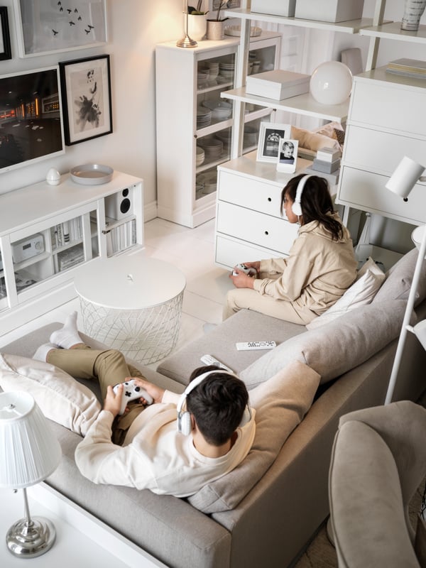 A living room in shades of white, with two teens in a beige FRIHETEN corner sofa-bed playing a video game, consoles in hand.