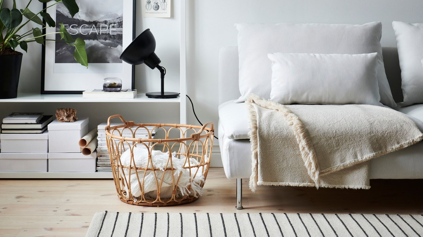Dans un salon aux couleurs claires, un canapé SÖDERHAMN blanc avec plaid beige, des étagères blanches avec boîtes de rangement, lampe de table noire, affiche noir et blanc et grande plante verte.