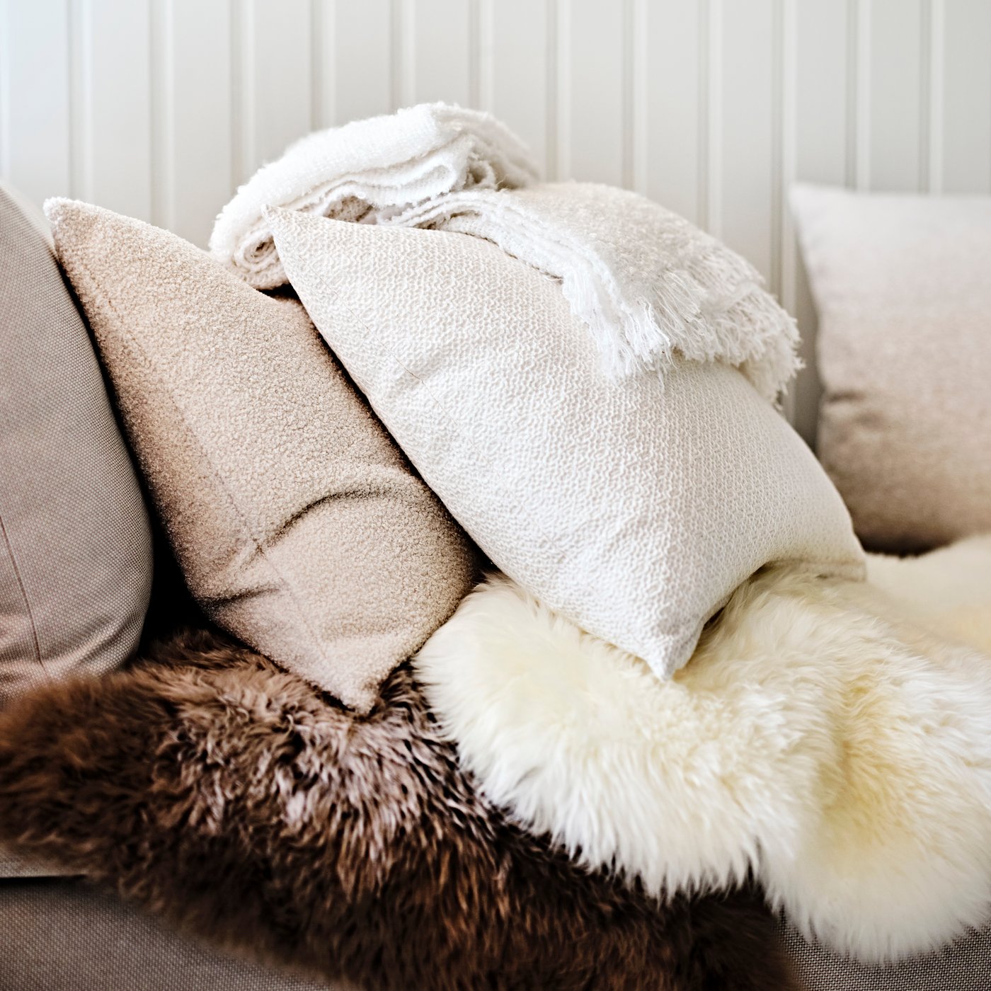 Novent inspiration i form av mjuka kuddar och filtar i beige och vitt ligger på en soffa med pälsplädar i brunt och vitt.