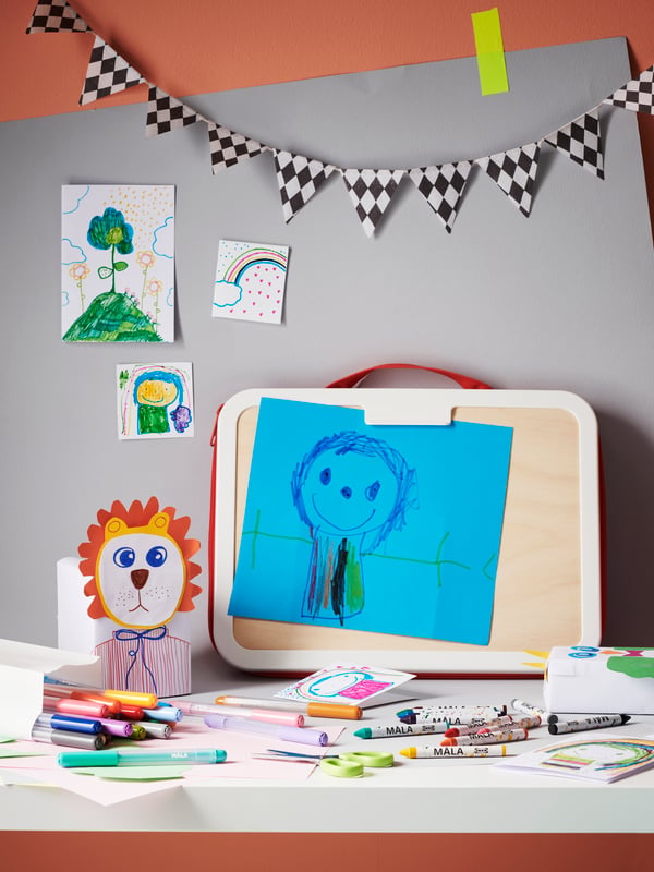 Un maletín para dibujar MÅLA apoyado sobre una mesa blanca y un muro cubierto por un cartón gris. Sobre la mesa hay lápices desordenados y sobre el muro dibujos infantiles.
