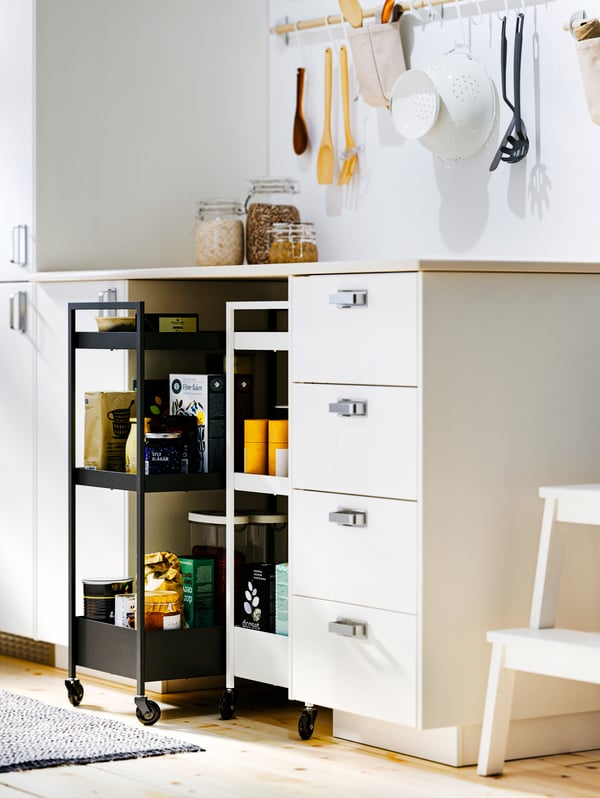 Dos carritos NISSAFORS, uno negro y uno blanco, ubicados bajo la encimera de un mueble de cocina de color blanco. Los carritos contienen utensilios y productos para cocinar. 