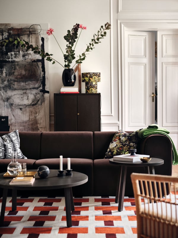 Dnevna soba u kojoj se nalazi tamnosmeđa STOCKHOLM 2025 sofa 4-sed, ispred koje su stolovi, a sve je smešteno na dezeniranom, ravno tkanom tepihu.