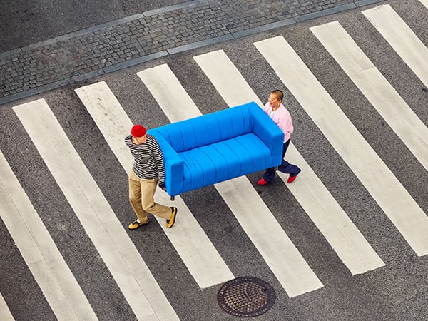 Two people carrying a blue KLIPPAN sofa whilst crossing a road.