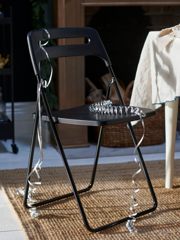 Silver streamers decorate a black NISSE folding chair set on a jute rug by a table. A fireplace is behind the chair.