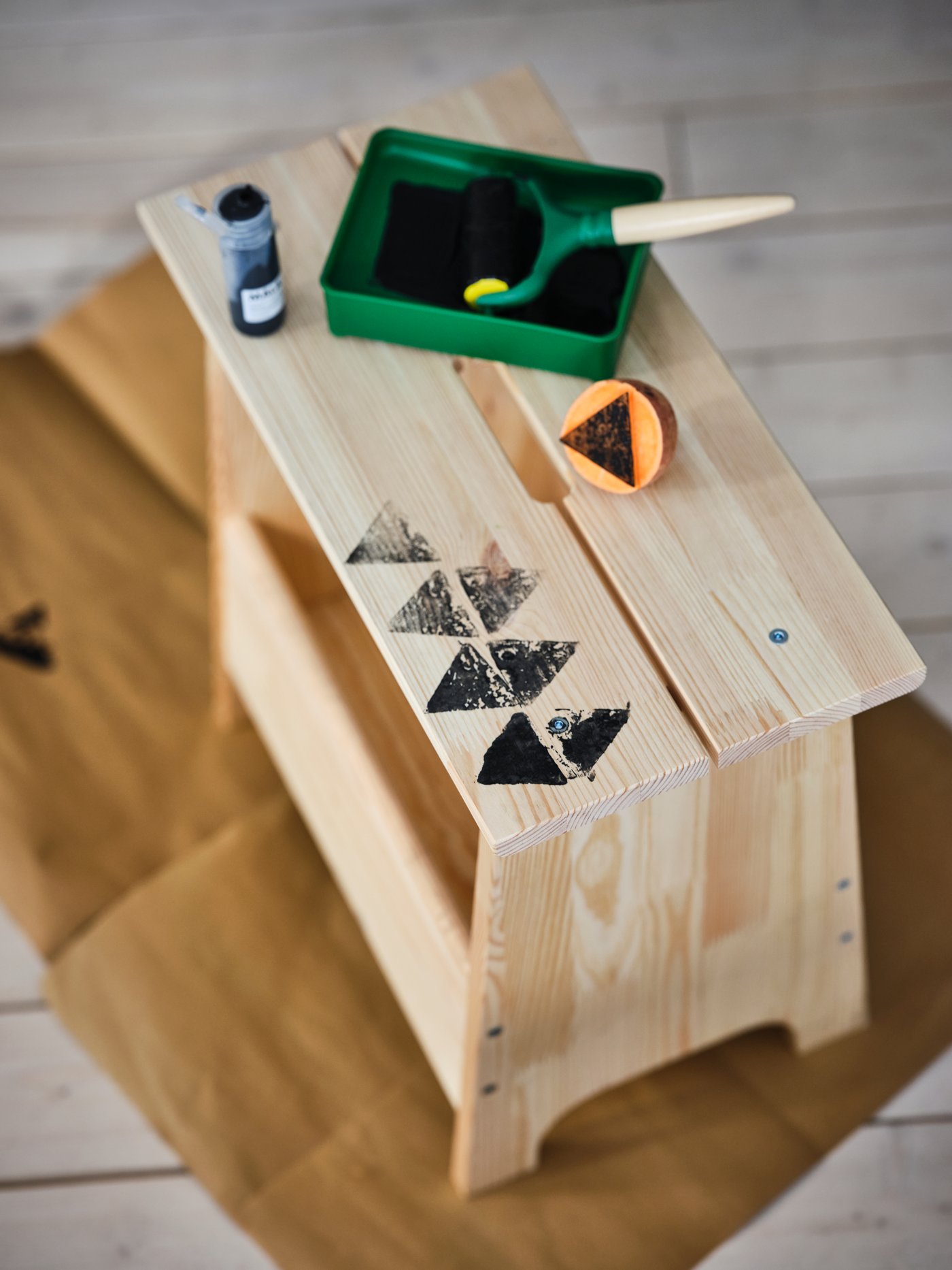 A child’s creative project takes shape on a pine IKEA PERJOHAN bench with storage, decorated with black triangle stamps and accompanied by a green tray containing a sponge and paint.