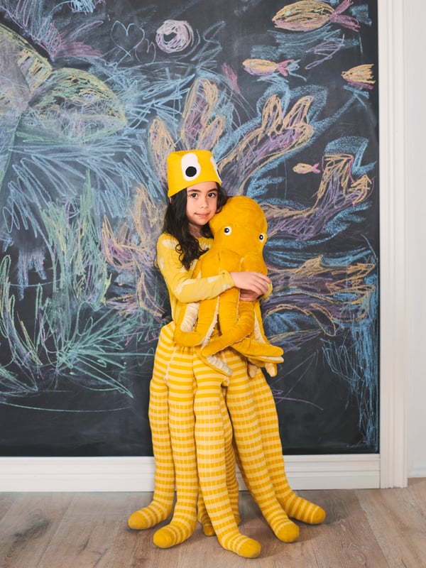 A young girl dressed in a handmade yellow octopus costume hugs a BLÅVINGAD octopus soft toy in front of a chalkboard wall.