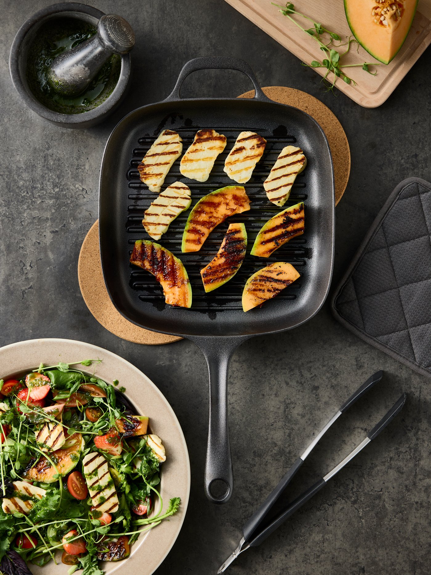 A VARDAGEN grill pan made of cast iron has grilled cheese and pieces of melon inside and there’s a salad bowl next to it.
