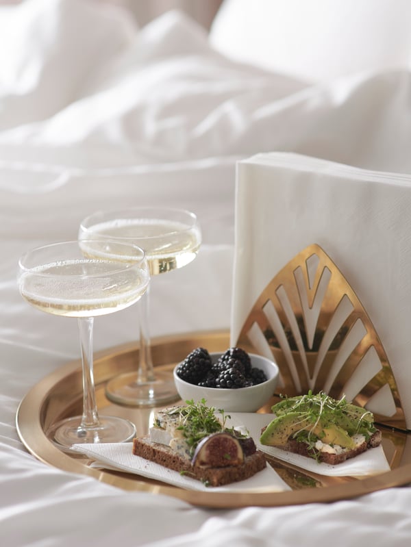 Copas STORHET rellenas de champaña en una bandeja en la cama junto a comida y un servilletero dorado TILLSTÄLLNING.