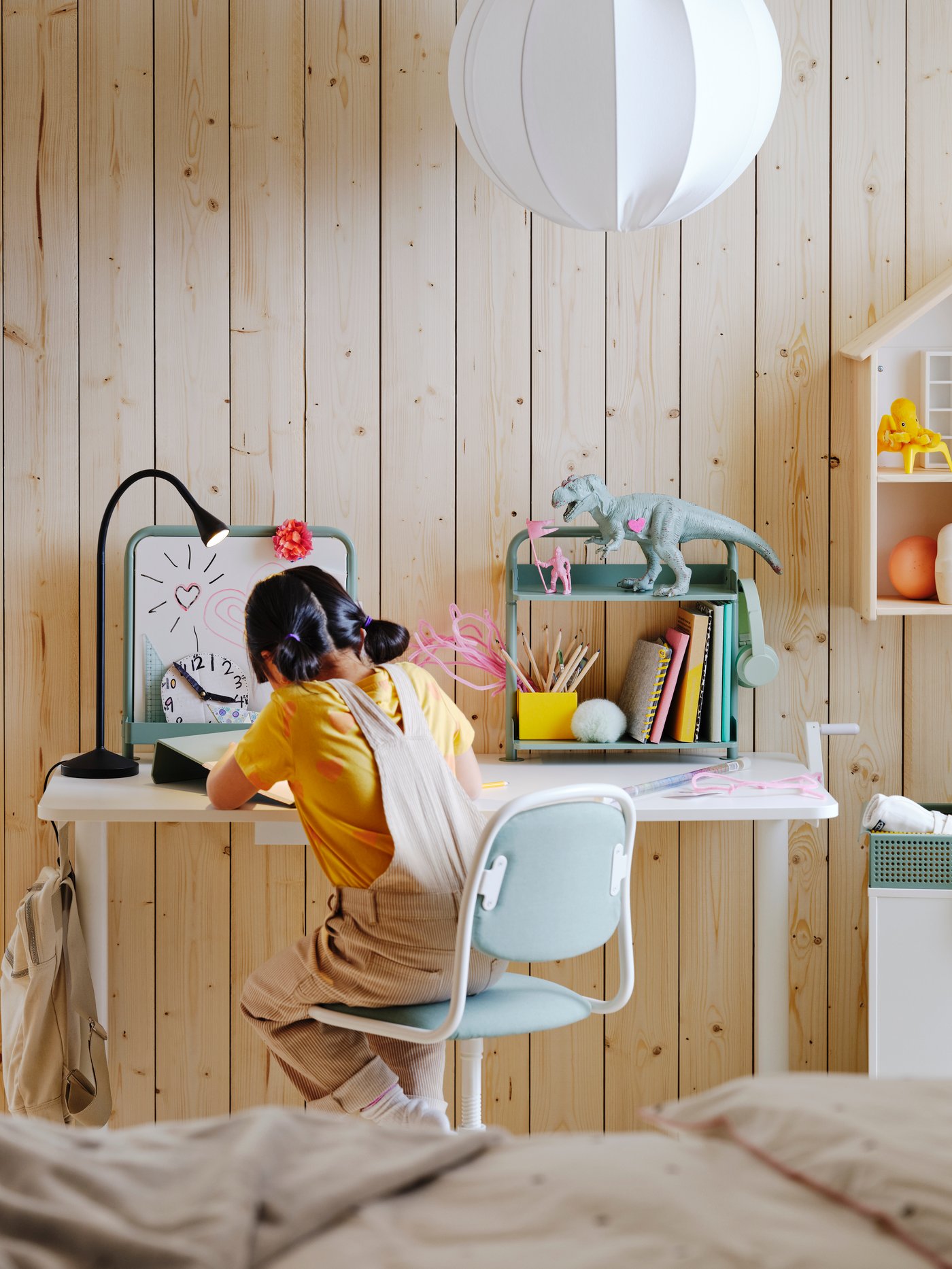 A child sitting on a green and white chair at a white RELATERA desk with a green bookshelf on it in a bright kid’s bedroom.
