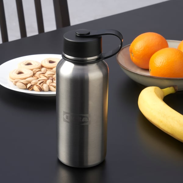 Silver insulated flask with black lid sits on a dark table, surrounded by snacks and fruit. The lid has a small loop for easy carrying.