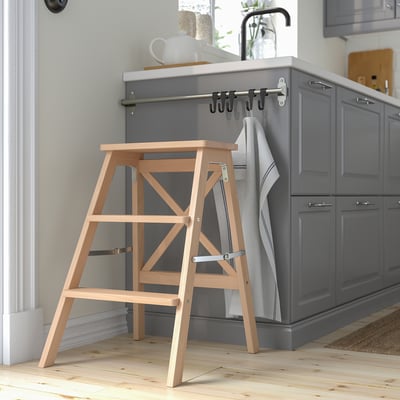 Wooden BEKVÄM step ladder against grey kitchen island. Foldable, light beech with three steps.