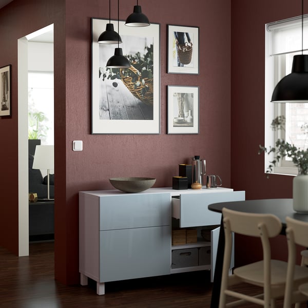 White BESTÅ sideboard with adjustable shelves and drawers. Pictures hang above, lights dangle from the ceiling. Wooden dining chair to the right.