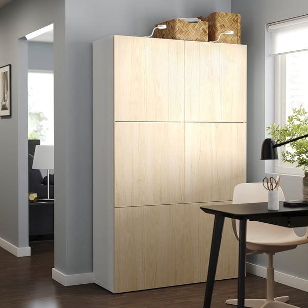 Image a light wood-colored BESTÅ storage unit in a modern living space, including a desk with a lamp and chair.