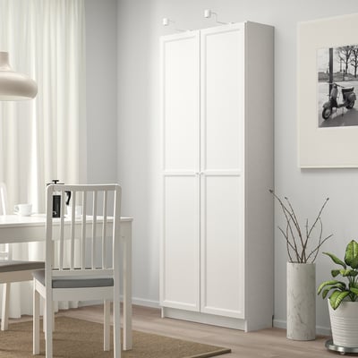 A modern, white BILLY bookcase stands tall, near a dining table and chair, offering ample storage. Its sleek design fits well in the bright room.