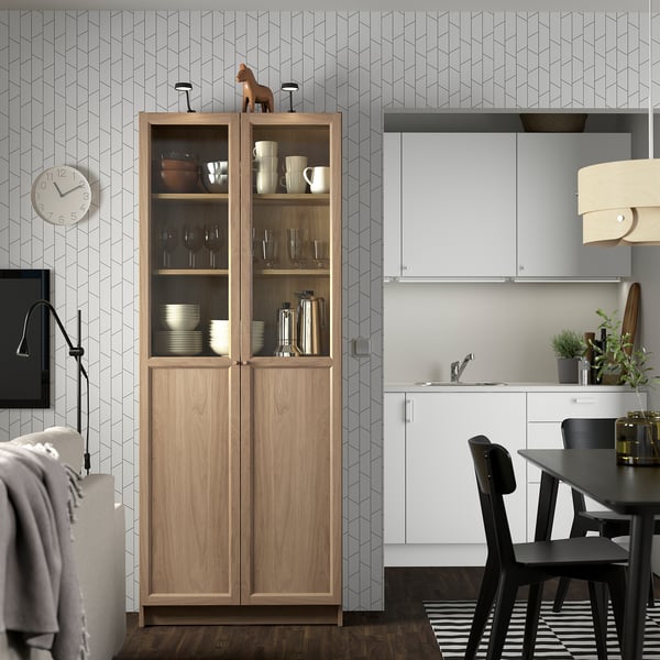 Bookcase with glass doors in modern kitchen. Wooden horse above, white sink, black chairs, wall clock, hanging lamp.