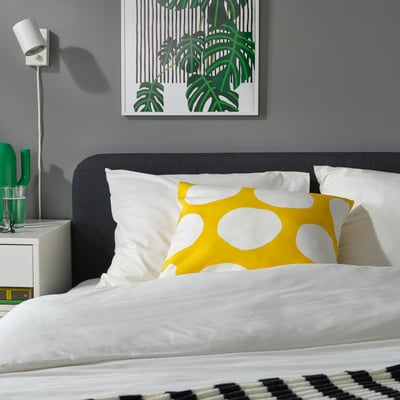 Neat bedroom with grey bed, white pillows, yellow BLÅMUNKAR dot cushion, green decor, and grey wall art.