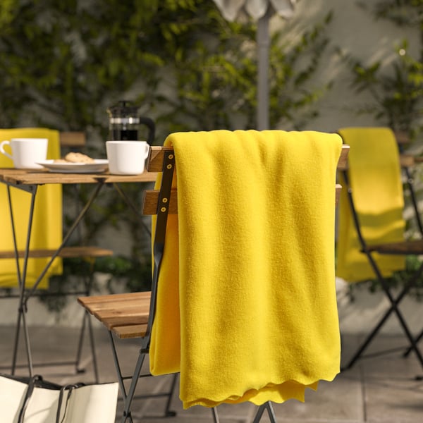 Bright yellow DAGGKÅPOR fleece blanket, soft & recycled, draped over chair, larger than cups on nearby table.