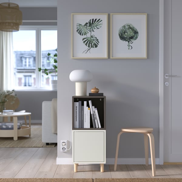 Modern living room with grey EKET shelves, books, lamp, prints, and wicker coffee table, natural light.