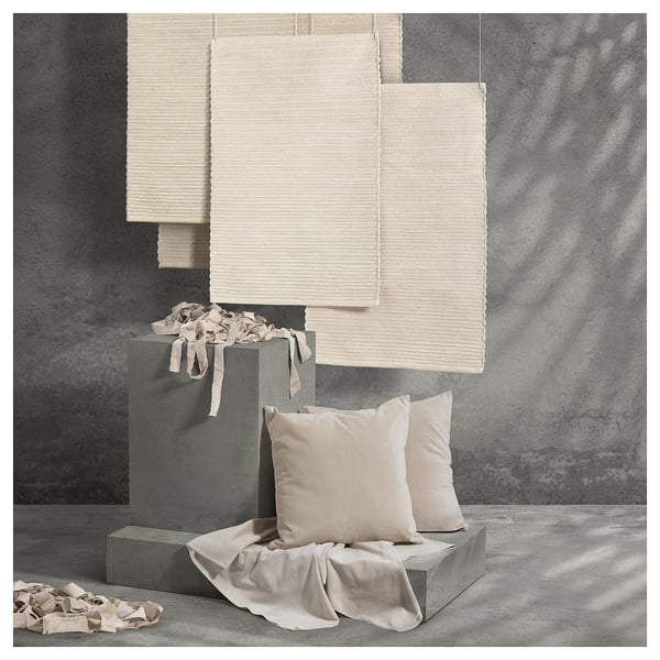ENERGIKÄLLA rug: handwoven, beige, cotton. Unique, square design. Set against grey wall and blocks, showcasing texture and craftsmanship.