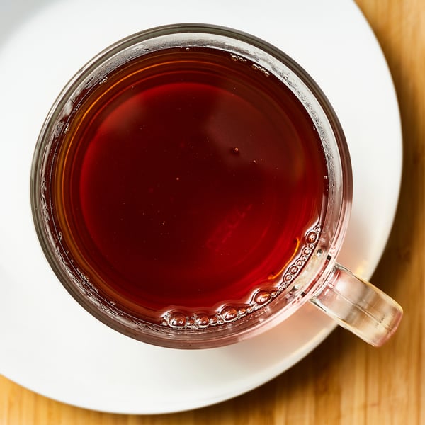 Clear glass mug filled with dark red tea on a white saucer. Calming, steaming beverage with rich flavour.