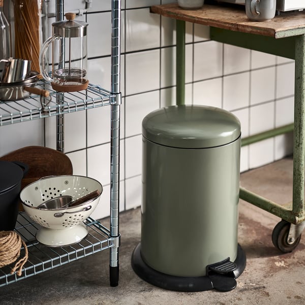 Olive green rubbish can with lid and foot pedal in kitchen next to metal shelf with items.