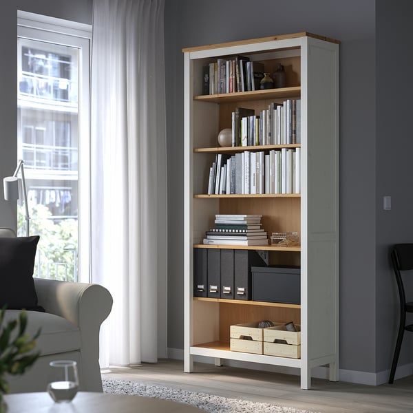 Tall, slim HEMNES bookshelf in white/pine, filled with books and decor, beside a window and grey sofa.
