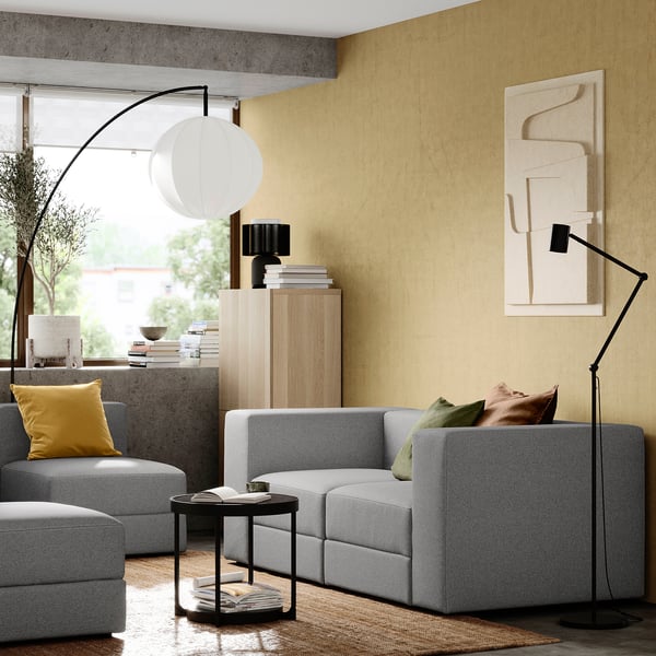 Modern living room with grey JÄTTEBO modular sofa, yellow pillows, floor lamp, side table, bookshelf with books, and abstract wall art.