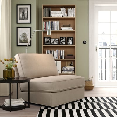 Modern living room with sage walls, a beige KIVIK sofa-bed, striped rug, and wooden bookshelf filled with books and black and white photos.