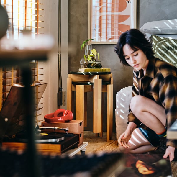 Person with baby on patterned sofa, wooden KOMPISHÄNG stools as plant stand, red toy on floor.